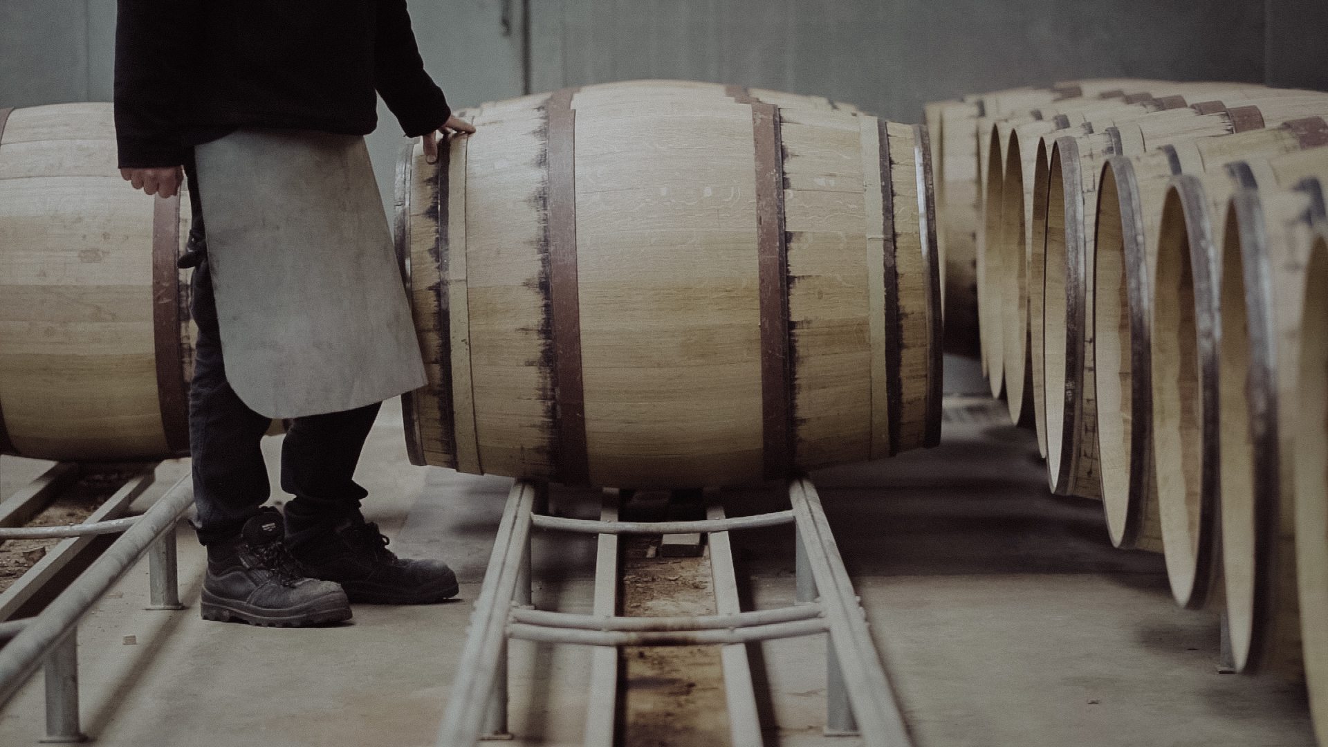 A cooper rolling a new oak barrel along the rails beside rows of finished barrels